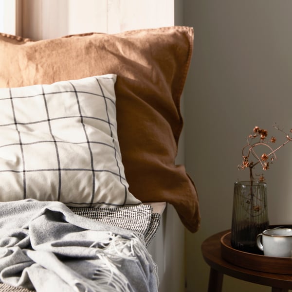 A cushion in an off-white/black SPIKKLUBBA cushion cover in front of another cushion in a dark beige DYTÅG cushion cover.