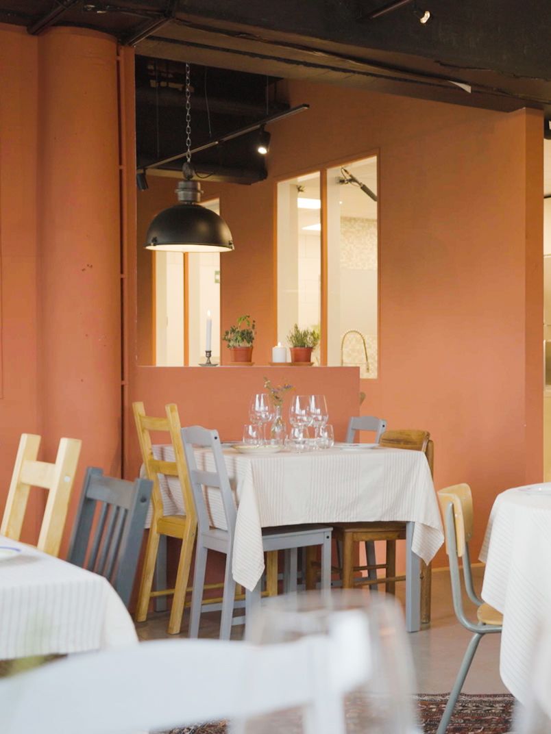Witte tafels met glazen en kleurrijke stoelen in warm interieur met terracottamuur. Planten op vensterbank en industriële lamp zorgen voor sfeer.