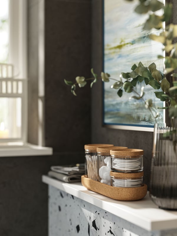 A set of five SAXBORGA jars with lids on a tray made of cork is standing next to a vase on a white cabinet in a brown room.