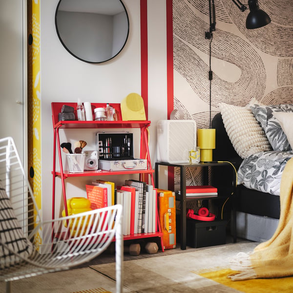 A red KRÖNIKÖR open storage unit holds books and beauty products against a stylish patterned wall by an upholstered bed.