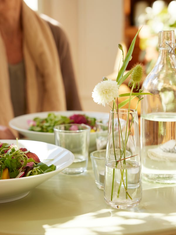 On a light and beautiful table setting with two white plates filled with sallad is a BERÄKNA glass vas with flowers.