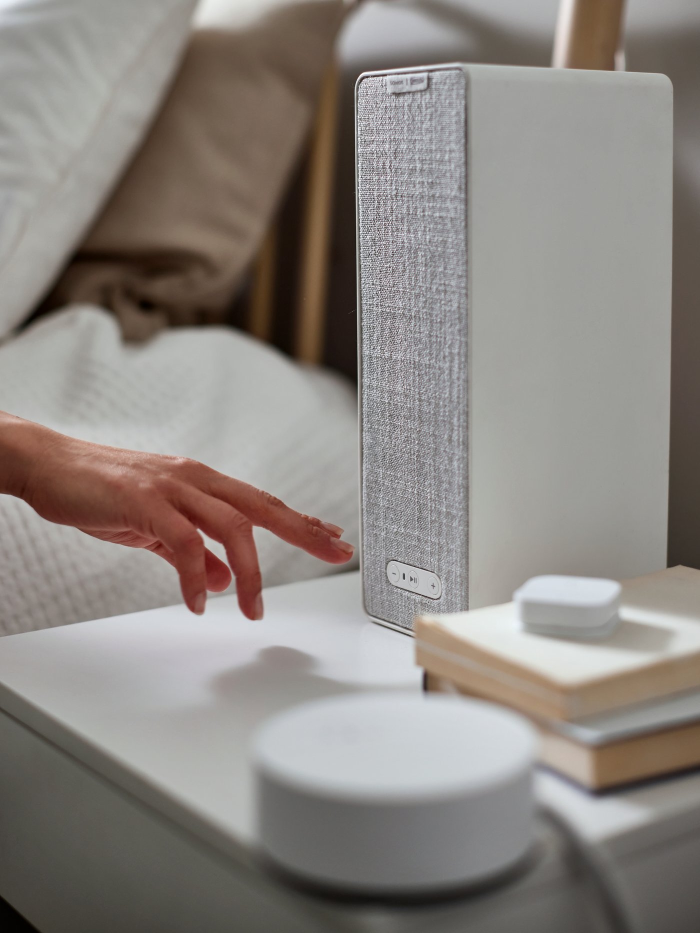 Una mano que presiona un control remoto que está sobre unos libros junto a un altavoz de estantería WiFi SYMFONISK en un buró blanco.