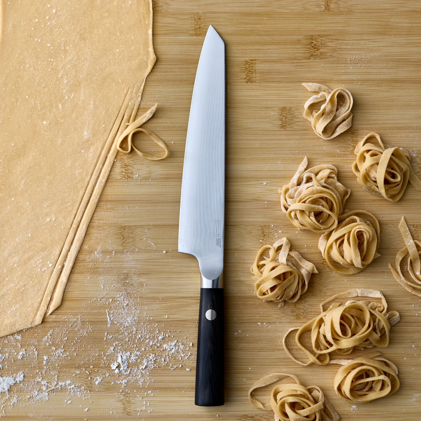 A wooden kitchen counter shows a close-up on a black FJÄLLRÖDING cook’s knife made from Damascus steel next to rolled pasta.
