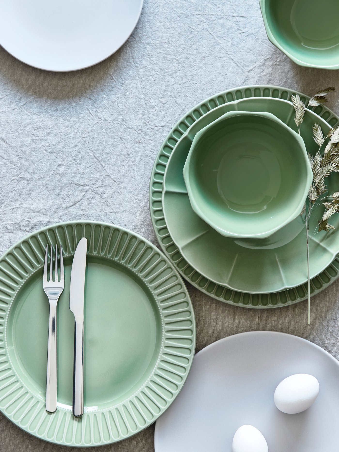 A table setting with STRIMMIG flower-inspired tableware in green. There is also a white plate with two eggs on the table.