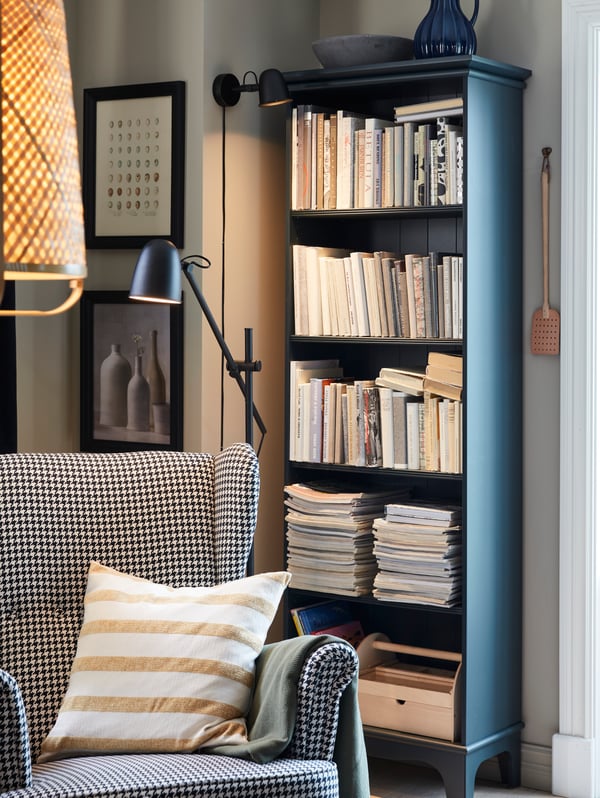 A cosy reading corner with a SKURUP wall lamp, a SKURUP floor lamp, a wing chair and a bookcase in dark blue-green.