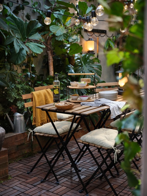 An outdoor table holding a serving tray with bowls of food, beside a kitchen towel, chairs with cushions and lamps.