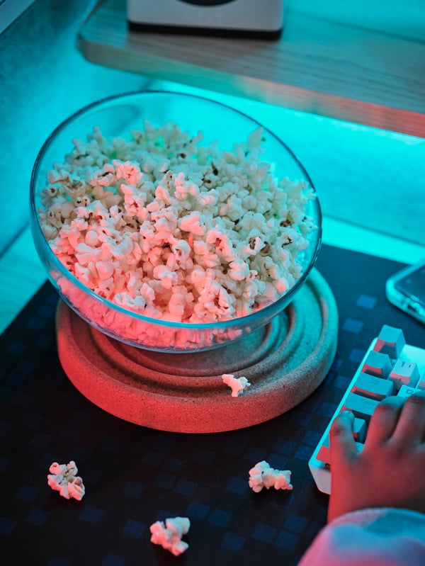 A BRÄNNBOLL coaster is set under a glass bowl full of popcorn on a desk. A BRÄNNBOLL gaming mouse pad is under the coaster.