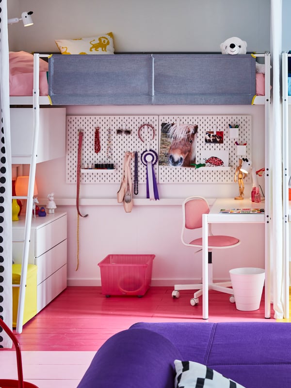 Barnrum med loftsäng, skrivbord och stol under. Pegboard med dekorationer på väggen, rosa golv, förvaring och del av lila soffa i förgrunden.