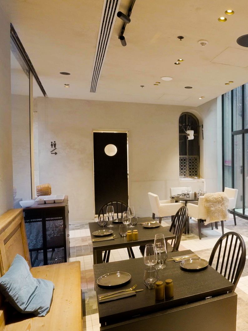 Modern restaurant met zwarte tafels, glazen en mix van stoelen. Houten bank met blauwe kussens en veel licht via grote ramen in strak interieur.