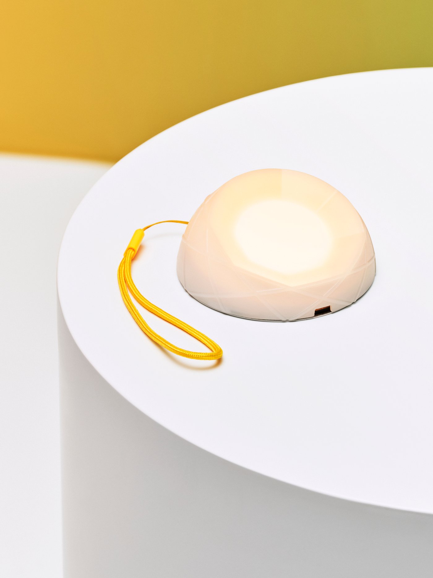 Pencahayaan berkuasa solar LED SAMMANLÄNKAD dengan tali pergelangan tangan berwarna kuning diletakkan di atas meja berwarna putih dengan dinding berwarna kuning di belakangnya.
