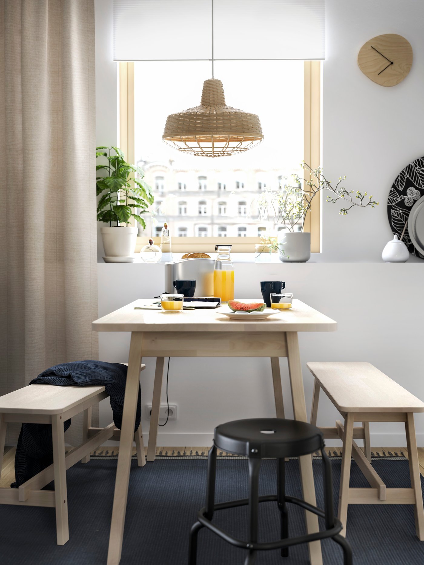 A dining spot next to a window with a wooden table, two benches, a black stool and a blue rug.