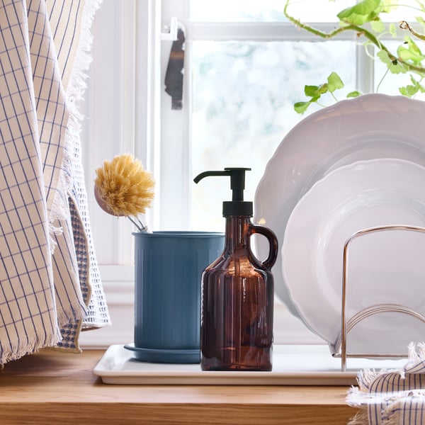 Plates are drying in a VÄLVÅRDAD dish drying rack and a dish brush is placed in a VÄLVÅRDAD cutlery stand on a worktop.