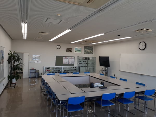 白い壁とホワイトボードに囲まれた会議室。青い椅子を並べたロの字型テーブルと天井の蛍光灯が見える。