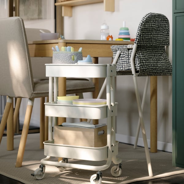 A white RÅSKOG trolley is placed next to a TONSTAD extendable table. It has baby-related items stored on the mesh shelves.
