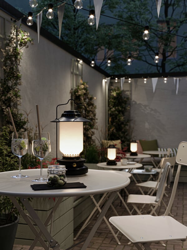 An outdoor cafe at night with black lanterns on beige outdoor tables, foldable chairs, lighting chains and bunting.