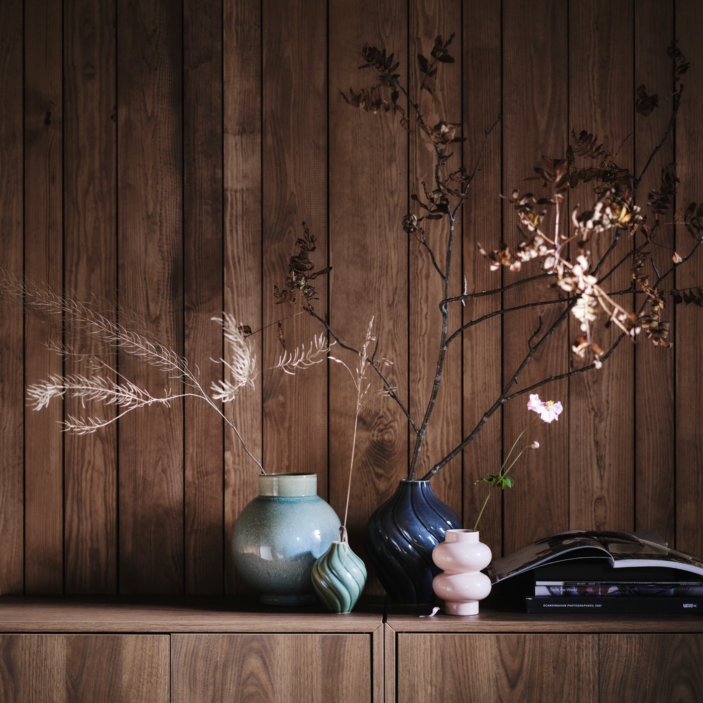 Four vases with branches stand on a wooden cabinet next to a stack of books. One of them is a light green GLANSGRAN vase.