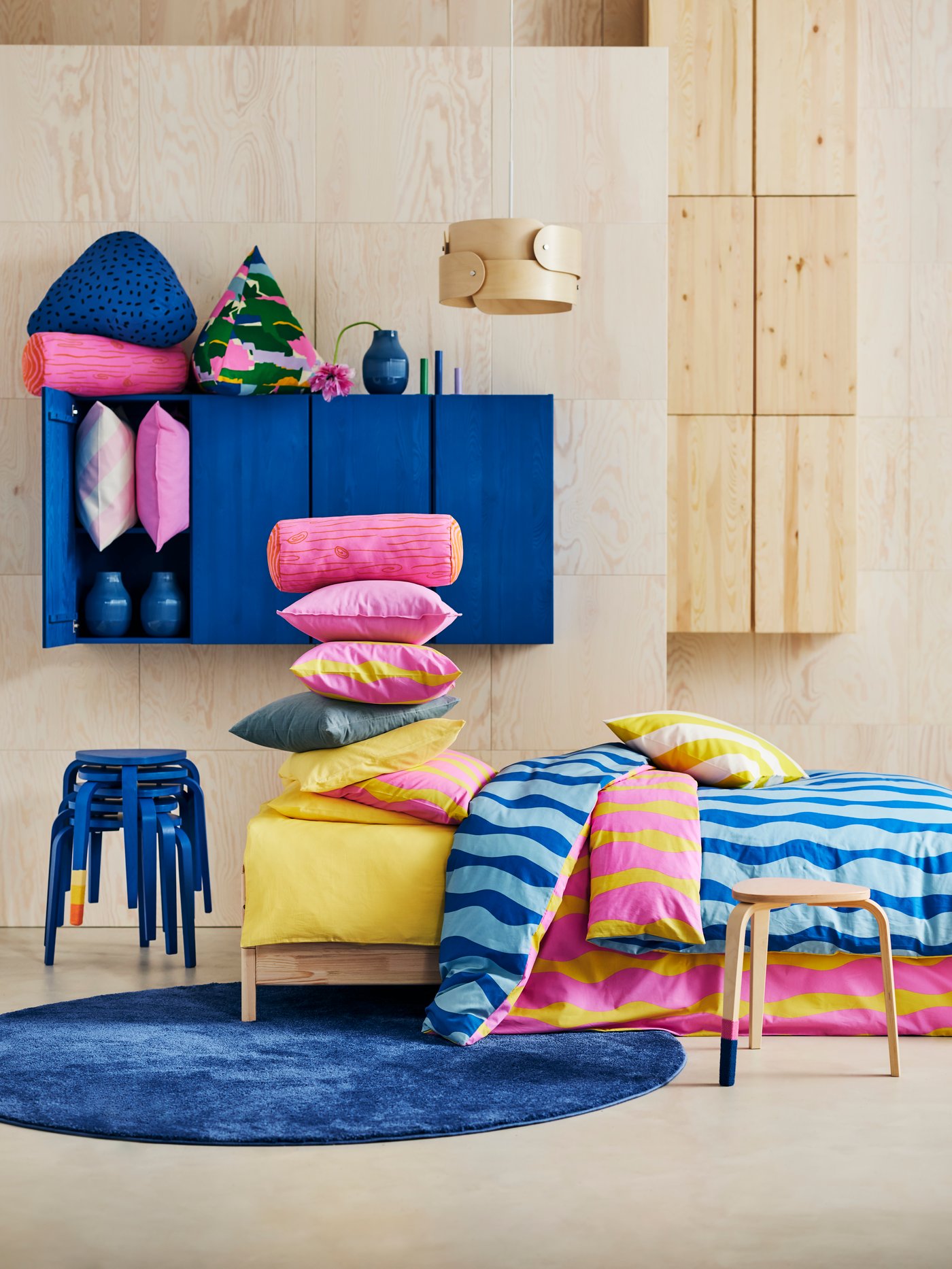 A bed with the SOMMARVICKER duvet cover and many pillows. Behind it, some KYRRE stools and some colourful EKLUGGMAL cushions.