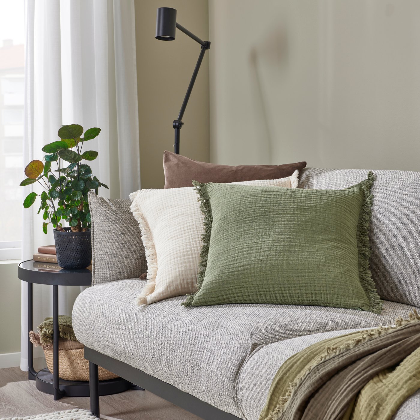 Auf einem hellgrauen Sofa befinden sich ein elfenbeinweißes und ein graugrünes VALLKRASSING Kissen aus 100% Baumwolle mit Knittereffekt und Fransen. Daneben liegen ein passendes Kissen aus braunem Samt und ein grünes Plaid.