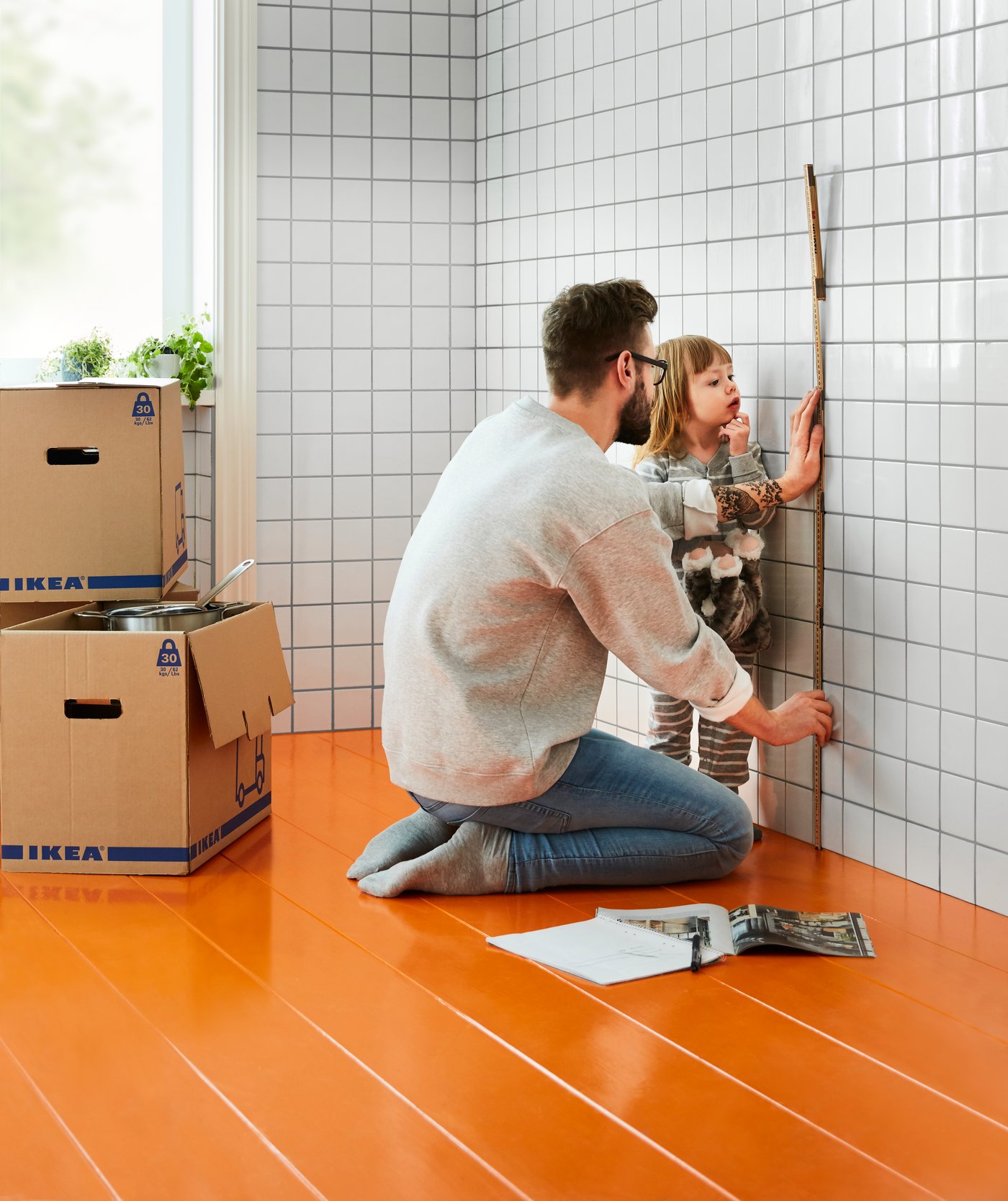 A man is crouching next to a girl in the room, measuring the room with a tape measure. Behind them are moving boxes.