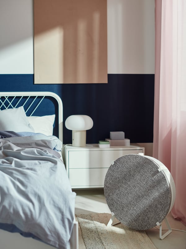 A black STARKVIND smart air purifier in front of a bookcase and a window that is covered in white/grey FJÄDERMOTT curtains.