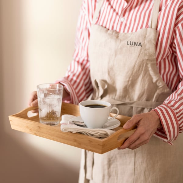 Una persona con un grembiule regge un vassoio FRUKTSKAL rettangolare in bambù, su cui sono appoggiati un bicchiere d'acqua con ghiaccio e una tazza di caffè.