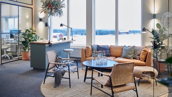 Modern kontorslounge med stora fönster som vetter mot ett snötäckt landskap. Inredningen inkluderar en ljusbrun lädersoffa med kuddar, två rottingstolar och ett runt soffbord med vattenflaskor och glas. Rummet är dekorerat med växter, inklusive en hängande växt ovanför en disk.