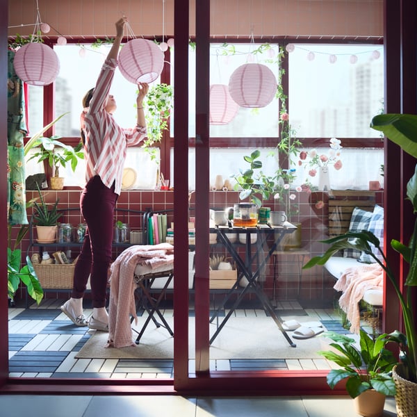 An enclosed balcony with a black and light brown TÄRNÖ outdoor table, surrounded by plants and decorative pink lamps.