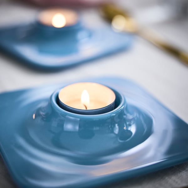 Two light blue VINTERFINT tealight holders have lit tealights in them, both are on a table.