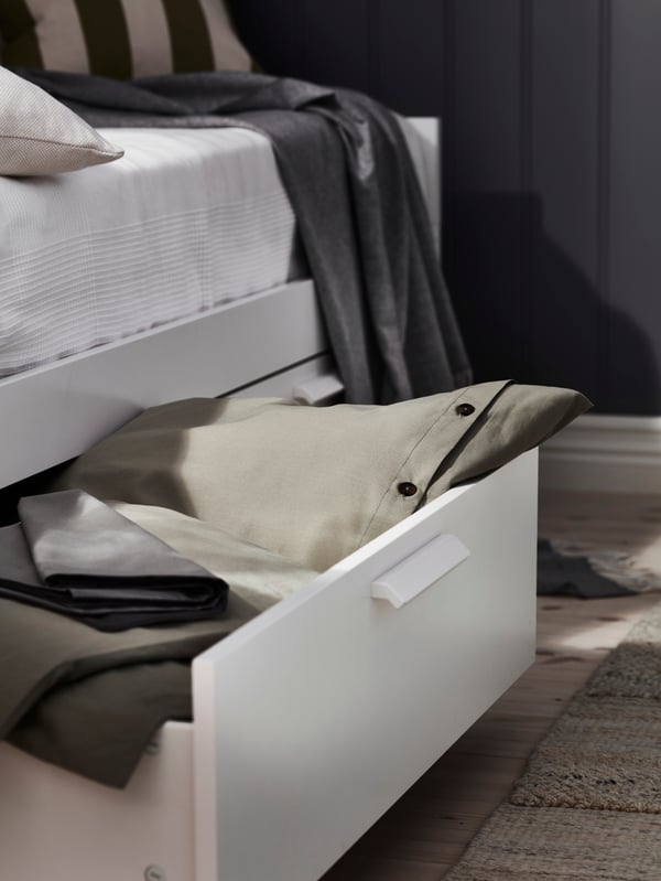 A drawer of a BRIMNES day-bed with 2 drawers has been pulled open. It contains bed linen, including KRÅKRISMOTT bed linen.