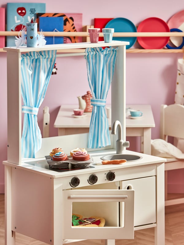 A SPISIG play kitchen with curtains stands in a child’s room with various toy food items on it and inside it.