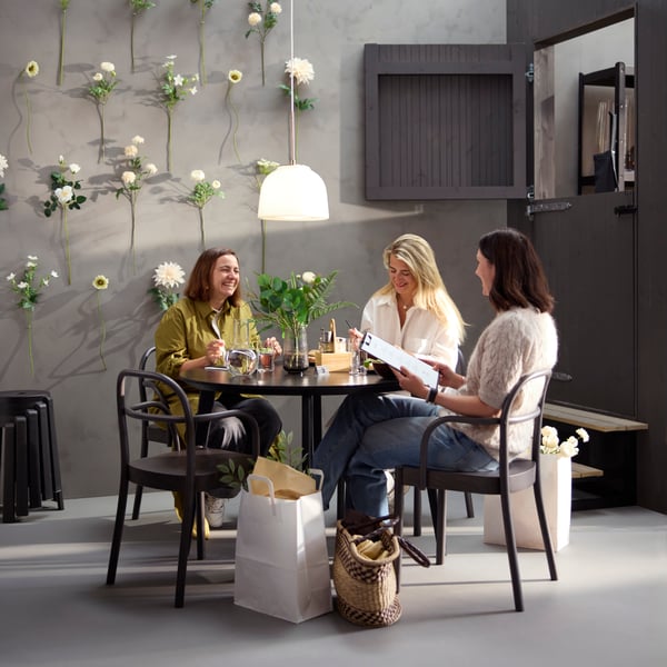 Café scene with three people at a black LISABO table, dark brown STOCKHOLM 2025 chairs, and a flower wall in the background.