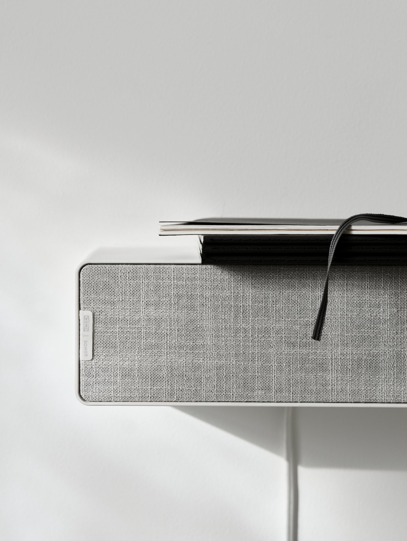 A white SYMFONISK WiFi bookshelf speaker attached to a wall with a small pile of books on top of it.