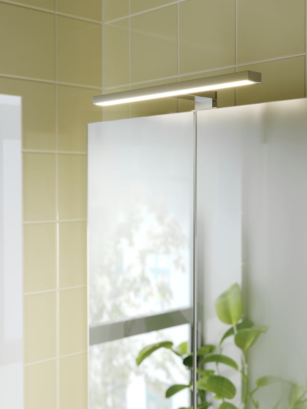 An aluminium coloured VÅTHULT LED cabinet mirror light is mounted above a bathroom cabinet on a wall with yellow tiles.