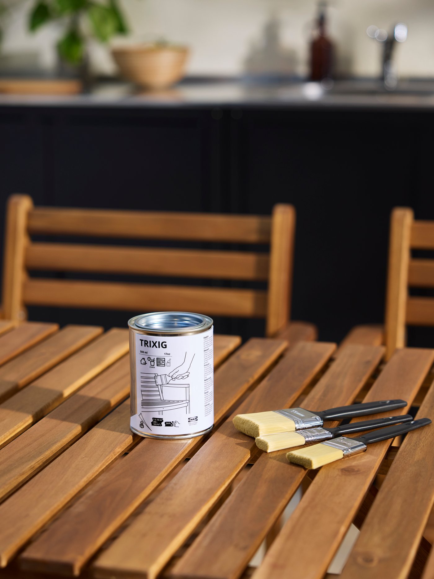 A wooden outdoor table with three brushes next to a can of TRIXIG outdoor wood stain placed on the table.