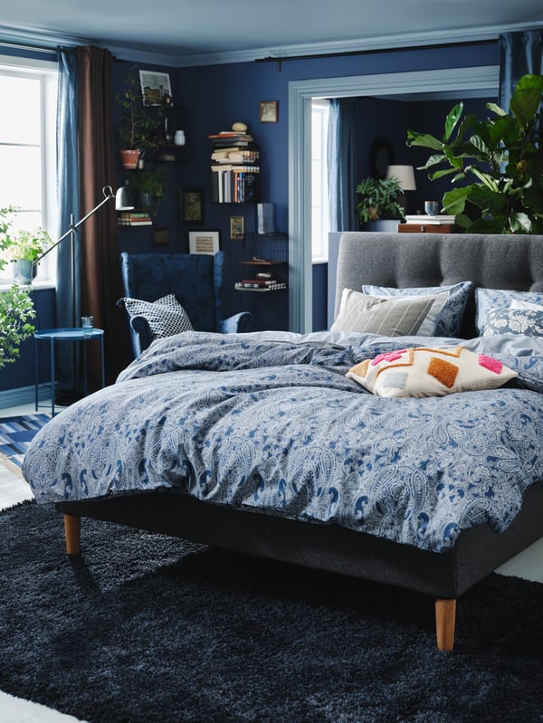 Cama gris IDANÄS con un cobertor JÄTTEVALLMO con diseños en color azul y blanco, en un dormitorio con tonalidades principalmente azules.