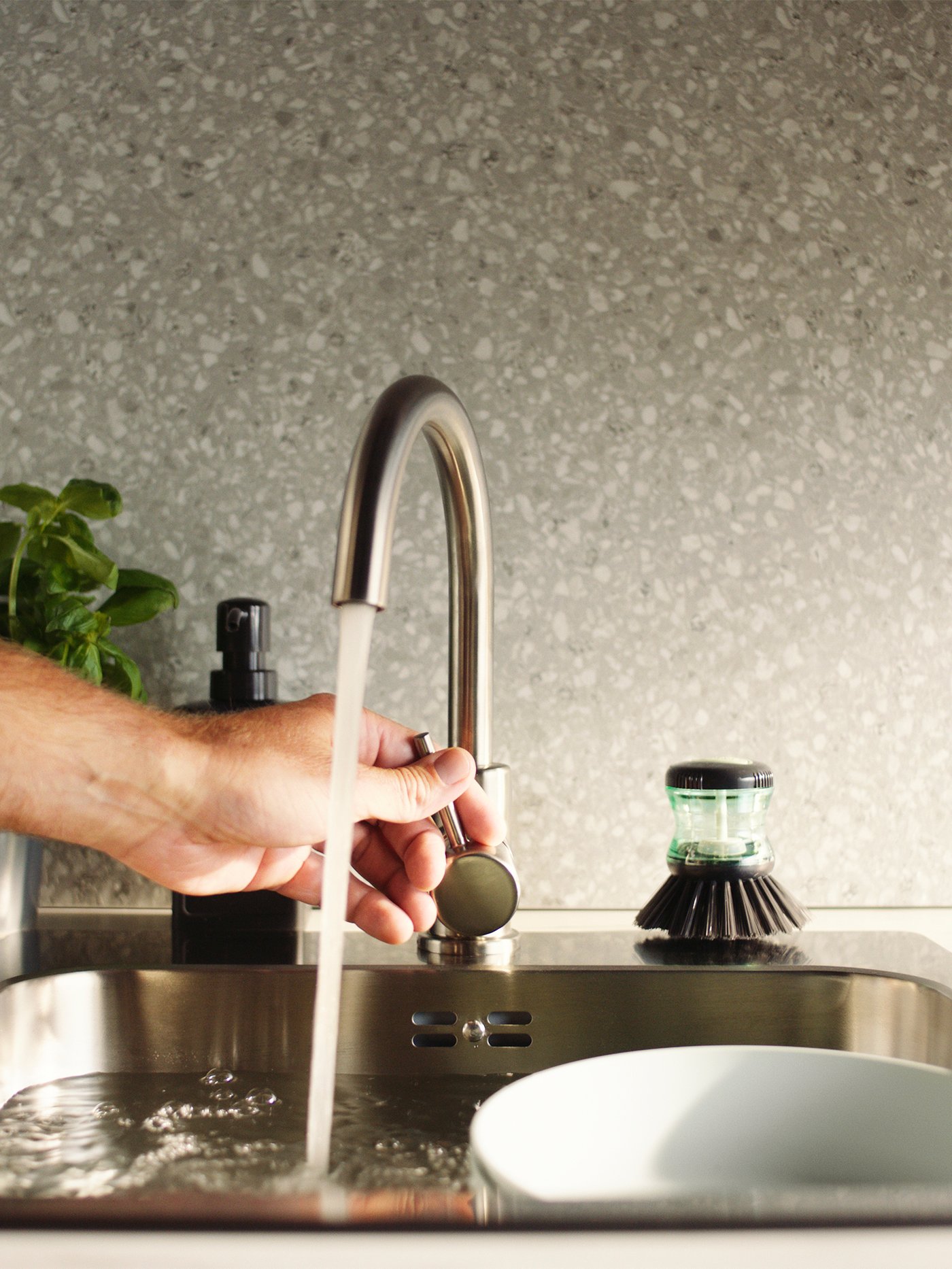 Hand adjusts the water flow of a GLYPEN kitchen tap in stainless steel next to a TÅRTSMET dish-washing brush.