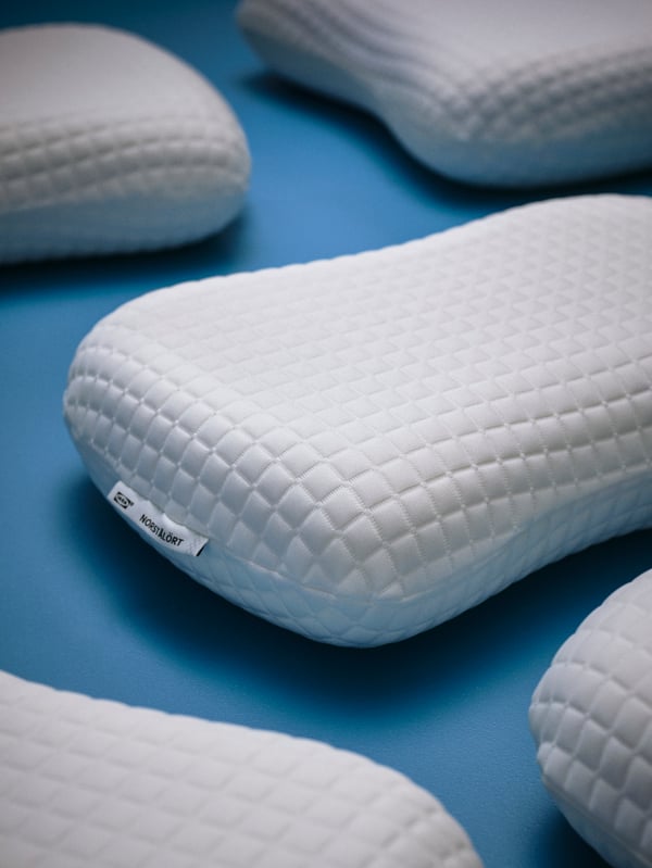 A white NORDSTÅLÖRT ergonomic pillow sitting on a blue surface, with four of the same pillows placed neatly around it.