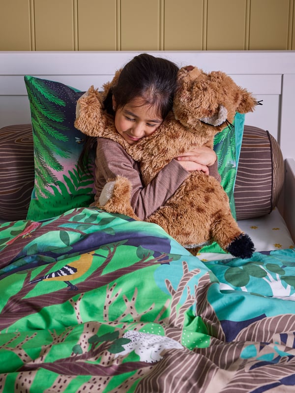 A child sits on a bed with SKOGSDUVA multi-coloured bed linen with a forest pattern and hugs a SKOGSDUVA lynx soft toy.