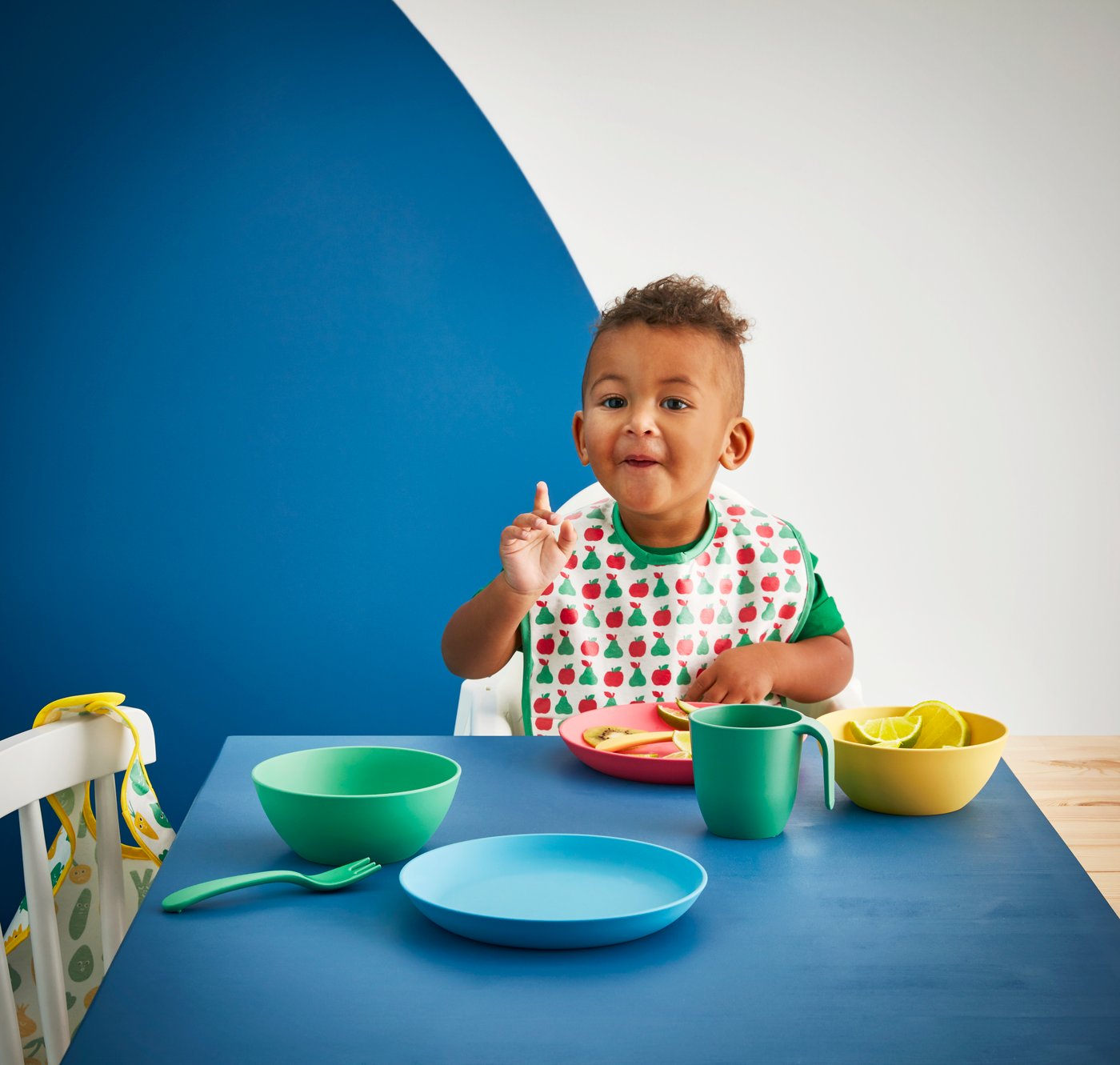A young boy wears a MATVRÅ Bib with patterns of fruits and vegetables, and sits in front of the dining table.