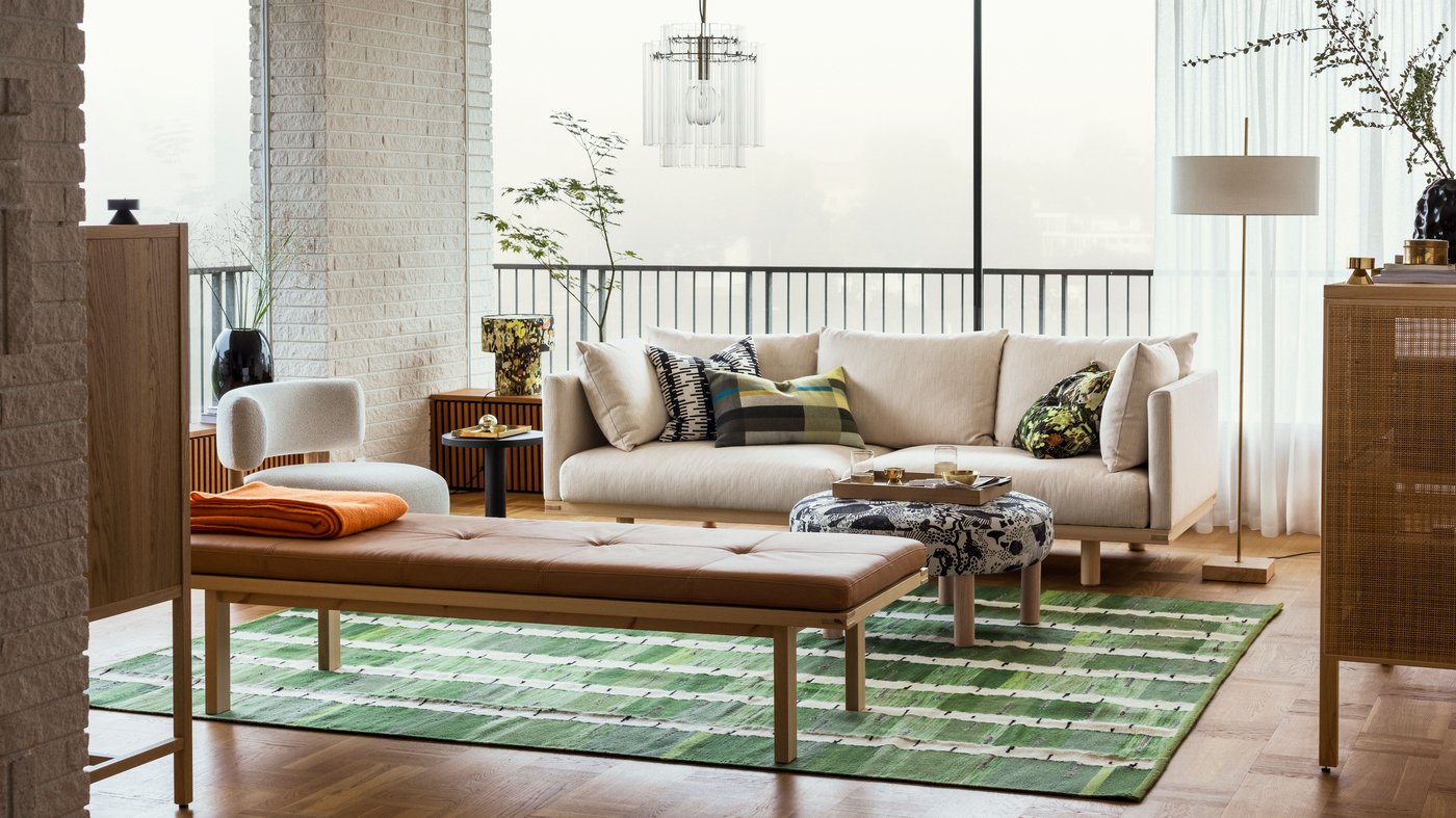 A bright living room with a beige STOCKHOLM 2025 3-seat sofa by a bench and pouffe sitting on a green, black, and white rug.