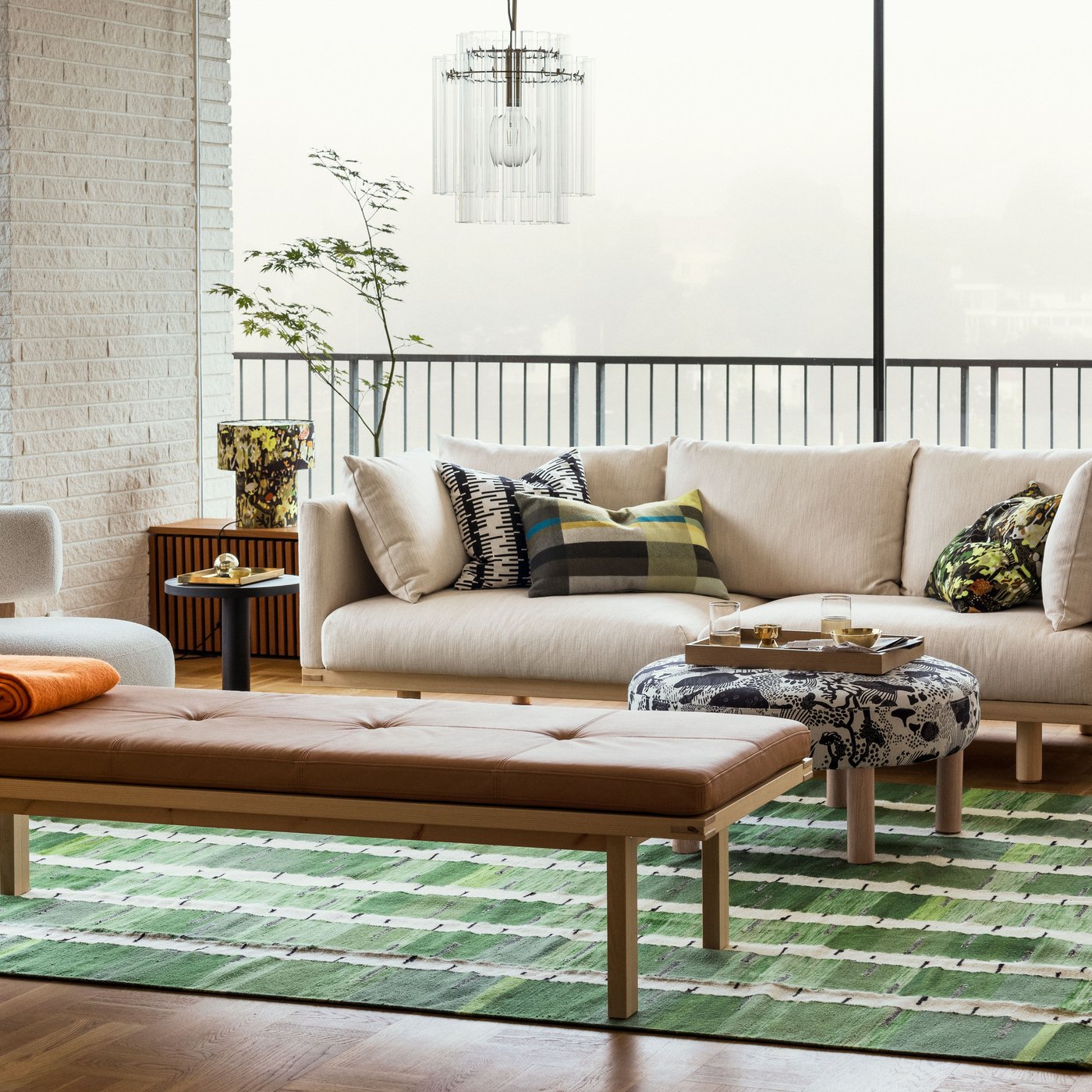 A bright living room with a beige STOCKHOLM 2025 3-seat sofa by a bench and pouffe sitting on a green, black, and white rug.