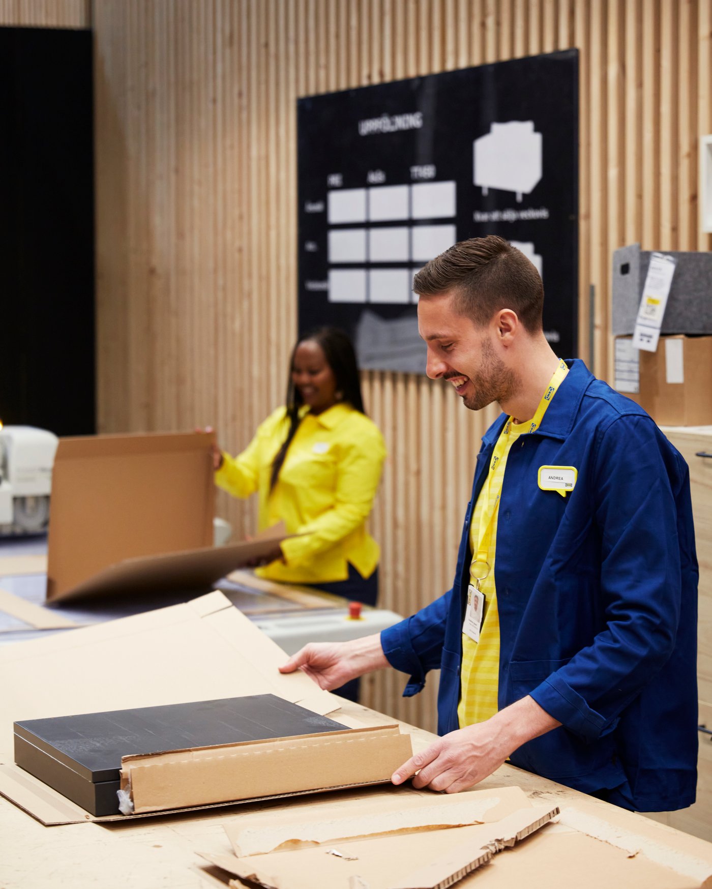 Twee IKEA medewerkers, een in een geel shirt en een in een blauw shirt, verpakken voorwerpen in karton.