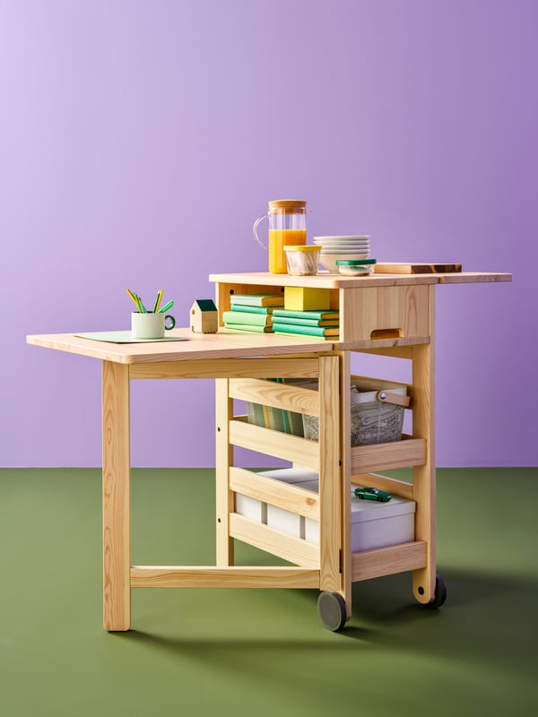 A RESARÖ mobile drop-leaf table with storage has boxes, baskets and books on the shelves and some other objects on the top.