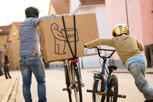 Eine erwachsene Person und ein Kind transportieren IKEA Pakete auf ihren Fahrrädern, die sie schieben.