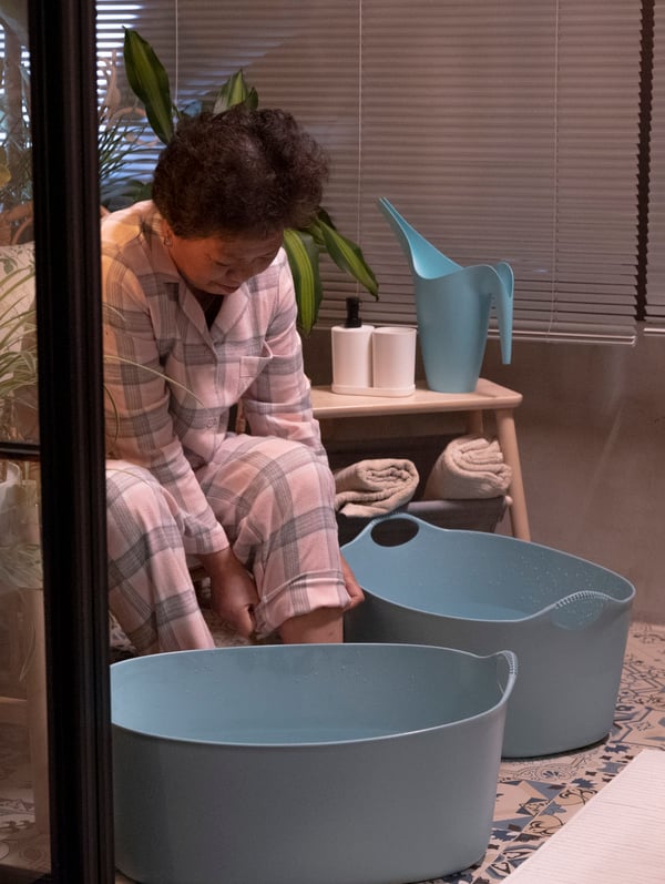 Woman getting ready for a cold and warm water foot bath, with two blue TORKIS flexi laundry baskets in front of her.