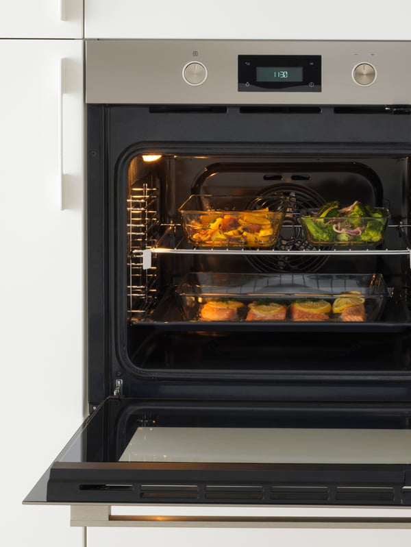An ANRÄTTA forced-air oven in stainless steel with the door open and three glass oven trays with different kinds of food.