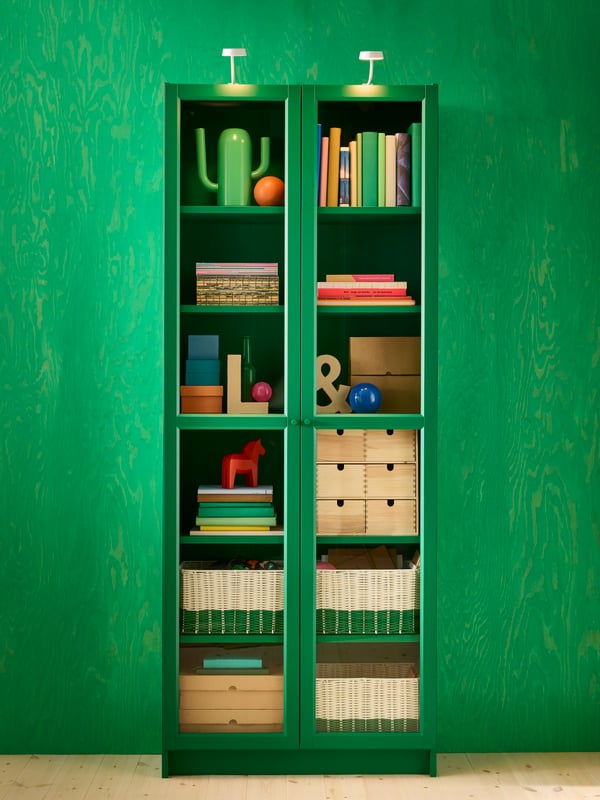 Tall green glass display cabinet filled with books and other items in front of green wall.