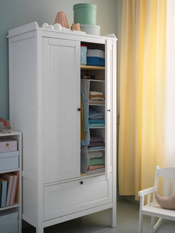 A SUNDVIK wardrobe stands by a window with a yellow curtain. One wardrobe door is open and there are clothes inside.