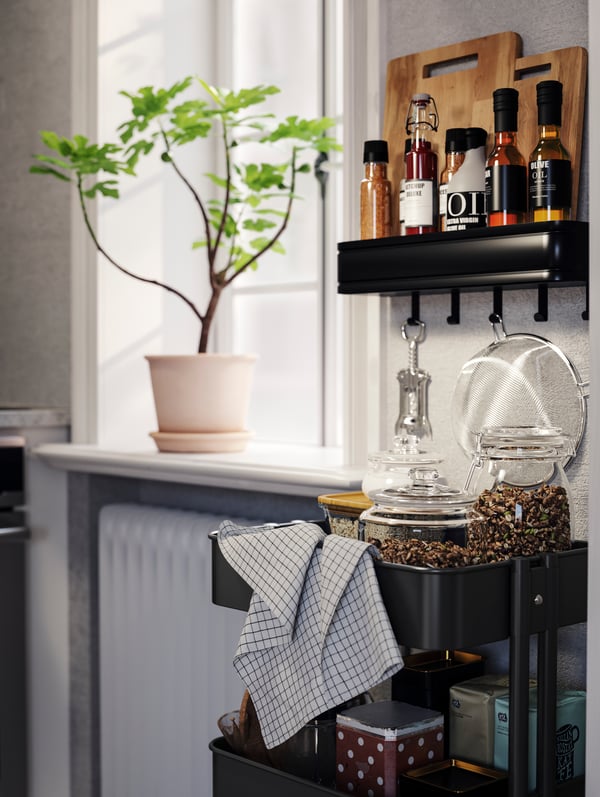 A RÅSKOG trolley with jars, a tea towel, and various kitchen items, placed next to a window with a potted plant on the sill.