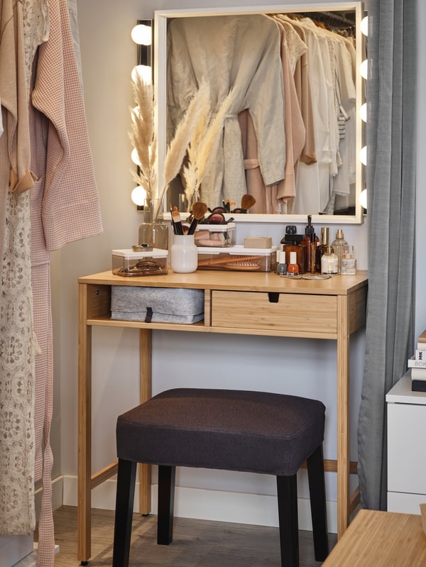 NORDKISA small wooden dressing table with an illuminated mirror and makeup products neatly organised on top. An upholstered stool stands in front of the table, with clothes visible in the mirror's reflection.